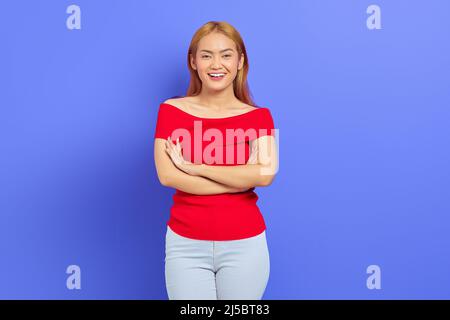 Ritratto di elegante donna asiatica in un bel vestito maroon in posa su fotocamera con le braccia ripiegate isolato su sfondo viola Foto Stock