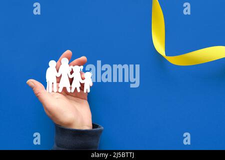 Aiuto mano che tiene ucraino famiglia, carta silhouette. Disposizione piatta, vista dall'alto. Spazio di copia, testo posizionato su sfondo blu con nastro giallo. Foto Stock