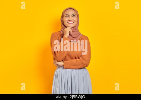 Bella giovane donna asiatica in maglione marrone e hijab sorridente guardando con mano sul mento, avendo buona idea isolato su sfondo giallo. Persone Foto Stock