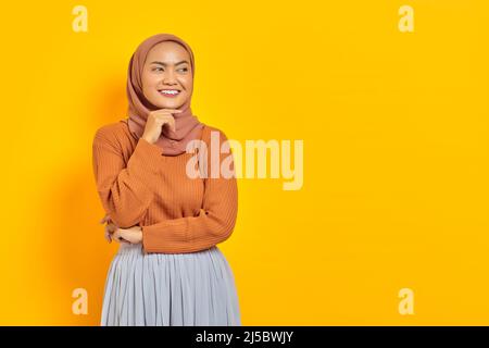 Bella giovane donna asiatica in maglione marrone e hijab sorridente guardando da parte con la mano sul mento, avendo buona idea isolato su sfondo giallo. Peop Foto Stock