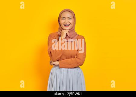 Bella giovane donna asiatica in maglione marrone e hijab sorridente guardando la macchina fotografica con mano sul mento, avendo buona idea isolato su sfondo giallo. Foto Stock