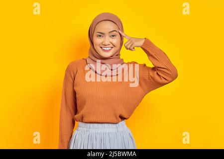 Bella giovane donna asiatica in maglione marrone e hijab sorridente guardando la macchina fotografica con il dito sulla testa, avendo grande idea isolato sopra giallo backg Foto Stock