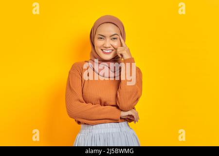 Bella giovane donna asiatica in maglione marrone e hijab sorridente guardando la macchina fotografica con il dito sulla testa, avendo grande idea isolato sopra giallo backg Foto Stock