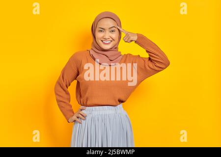 Bella giovane donna asiatica in maglione marrone e hijab sorridente guardando la macchina fotografica con il dito sulla testa, avendo grande idea isolato sopra giallo backg Foto Stock