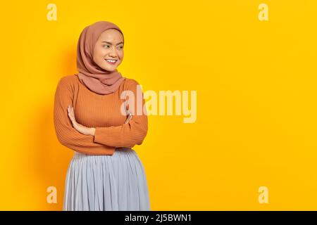 Bella donna asiatica sorridente in maglione marrone e hijab incrociato braccia petto e guardando lo spazio copia isolato su sfondo giallo. Persone islam re Foto Stock