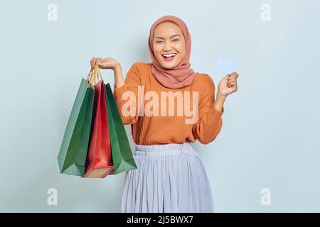 Eccitata bella donna musulmana asiatica in occhiali da sole, maglione marrone e hijab mostrando carta di credito, tenendo in mano shopping bag con acquisti dopo lo shopping è Foto Stock