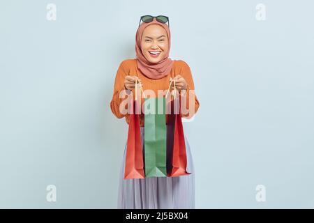 Eccitata bella donna musulmana asiatica in maglione marrone e hijab, tenendo in mano la borsa con acquisti dopo lo shopping isolato su sfondo bianco. Mondo Foto Stock