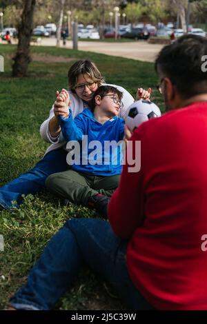 Ragazzo con disabilità ama giocare con una palla di calcio con i suoi genitori all'aperto in un parco. Foto Stock