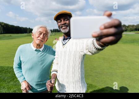 felice uomo afroamericano che prende selfie con un amico asiatico su smartphone blurred Foto Stock