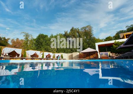 Area piscina con gazebo, sedie a sdraio e ombrelloni lungo la piscina nel resort di lusso. Case estive circondate da verde e blu Foto Stock