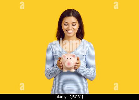 Gioiosa giovane donna casual che tiene la banca del piggy in forma di maiale isolato su sfondo giallo. Foto Stock