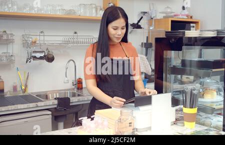 Giovane donna che è proprietario di affari della caffetteria che indossa un grembiule in piedi da un terminale del punto vendita mentre lavora nel caffè moderno. Foto Stock