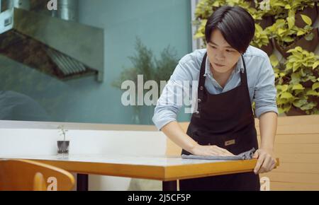 Una dipendente di un ristorante femminile o un giovane imprenditore pulisce il tavolo da pranzo in preparazione per accogliere i clienti. Foto Stock