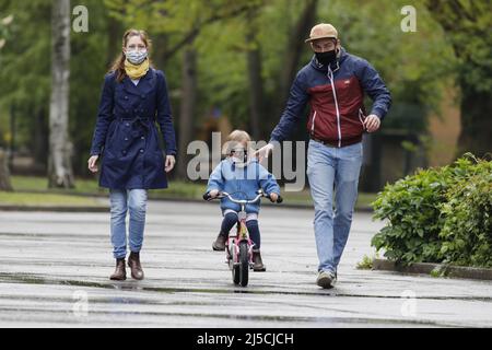 Finalmente uscire - la famiglia indossa maschere facciali in un parco pubblico mentre si pratica in bicicletta con la loro figlia giovane. [traduzione automatizzata] Foto Stock