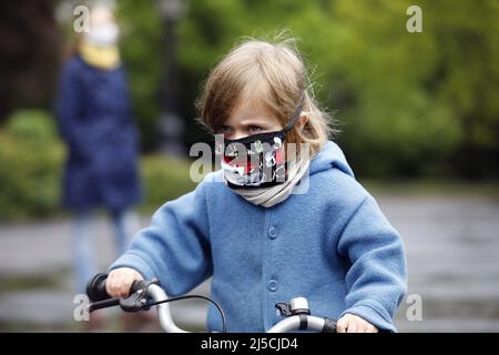 Finalmente uscire - la famiglia indossa maschere facciali in un parco pubblico mentre si pratica in bicicletta con la loro figlia giovane. [traduzione automatizzata] Foto Stock