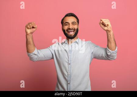Fortunato. Ragazzo indiano eccitato che celebra la vittoria e gesturing sì con i pugni su sfondo rosa. Uomo con portico multirazziale in jeans casual maglia vincente Foto Stock