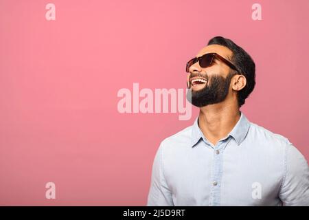 Uomo indiano allegro in occhiali da sole ridendo toothy, spensierato ragazzo ispanico in maglietta blu in piedi e ride fuori forte su sfondo rosa. Stile di vita felice Foto Stock