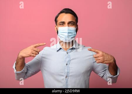 Proteggersi durante la pandemia. Il giovane indiano punta le dita sulla maschera medica sul suo volto isolato su rosa. Salvataggio dal concetto allergico, smog, virus Foto Stock