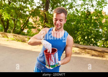 Il ragazzo dipinge il muro con un pennello in colori luminosi, tiene una lattina di vernice in mano, sulla strada Foto Stock