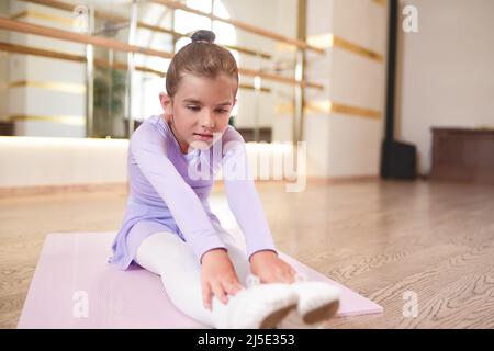 Adorabile foto di ballerina in abito lilla e collant bianco esercizi di stretching in una sala con grandi specchi. Foto Stock