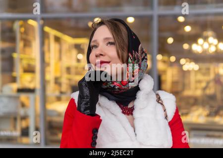 giovane donna sognante felice in un foulard in clima freddo si erge all'esterno sullo sfondo di un blurry luminoso vetrina Foto Stock