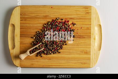 Una miscela di peperoni multicolore. Spezie aromatiche per alimenti. Fondo di semi di pepe secchi su un asse da cucina di legno con un cucchiaio graduato. Foto Stock