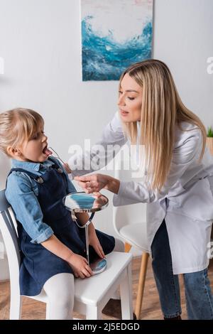 Il terapista di discorso in cappotto bianco che tiene lo strumento vicino alla ragazza e specchio in aula Foto Stock