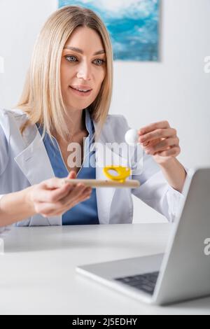 Logopedist positivo che tiene trainer del muscolo respiratorio durante la videochiamata sul laptop nella stanza di consulenza Foto Stock