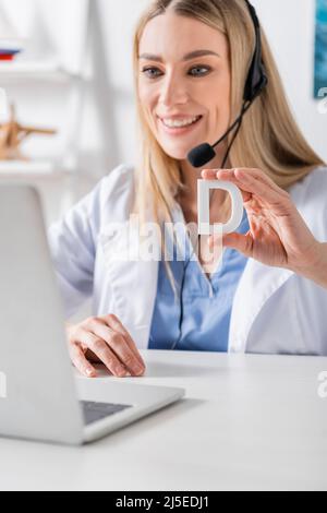 Logopedist sorridente in cuffia che tiene la lettera vicino al laptop durante la videochiamata nella stanza di consulenza Foto Stock