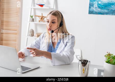 Terapista vocale in cuffia con video chat e puntamento con le dita sul laptop in sala di consulenza Foto Stock