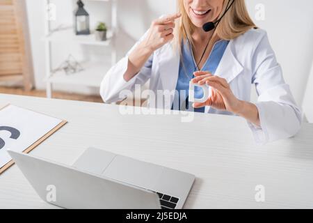 Vista ritagliata di un logopedist sorridente in cuffia che tiene la lettera e punta con le dita durante la videochiamata sul laptop nella stanza di consulenza Foto Stock