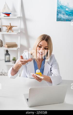 Logopedist sorridente che tiene Trainer del muscolo respiratorio mentre parla durante la videochiamata sul laptop nella stanza di consulenza Foto Stock