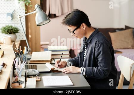 Concentrato giovane studente asiatico in occhiali seduti al tavolo in sala campus e disegno in sketchpad Foto Stock