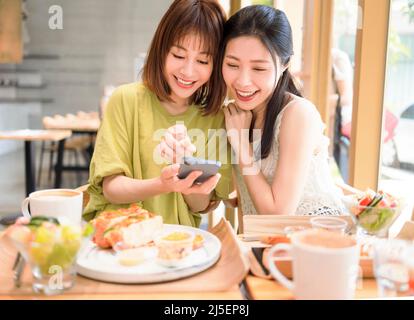 Giovani donne seduti al ristorante guardando il telefono cellulare e sorridenti Foto Stock