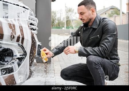 Vista laterale di ragazzo serio ed elegante seduto accanto alla sua automobile in auto lavaggio, utilizzando spugna saponata per schiumare e pulire la targa sulla macchina. Foto Stock