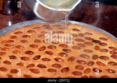 Versando lo sciroppo zuccherino di miele caldo su un vassoio di basbosa con mandorla , la Basbuda è un dessert mediorientale che viene solitamente cotto nel mese del Ramadan, selectiv Foto Stock