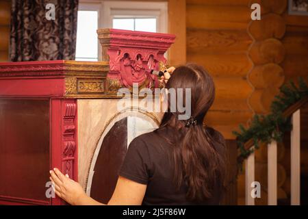 Donna colorando ordinatamente la parte superiore di armadio in legno arrugginito con splendidi ornamenti di colore rosso con pennello. Riutilizzo del vecchio. Laboratorio di antiquariato antico Foto Stock