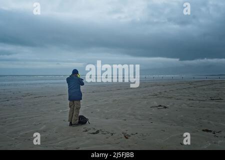 Uomo che scatta una foto a Tirella Beach, Irlanda del Nord, in un pomeriggio invernale sovrastato, guardando in lontananza le Mourne Mountains. Foto Stock