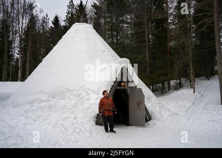 Rovaniemi, Finlandia - 18th marzo 2022: Un uomo sami in abiti tradizionali, in piedi alla porta della sua casa di legno sami coperto di neve tradizionale, chiamata Foto Stock