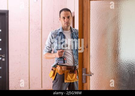 Operatore che installa o ripara la nuova serratura e la nuova manopola della porta con un cacciavite sulla vecchia porta. Foto Stock