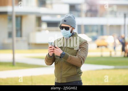 Un uomo in una maschera facciale indossa occhiali da sole che tengono uno smartphone su una strada accogliente. Foto Stock