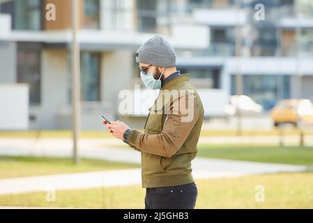 Un uomo in una maschera facciale indossa occhiali da sole che tengono uno smartphone su una strada accogliente. Foto Stock