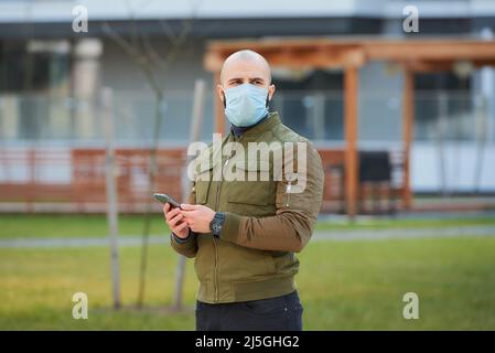 Un uomo calvo in una maschera facciale che controlla il suo smartphone su una strada accogliente. Foto Stock