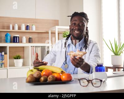 Nutrizionista Afroamericano che guarda la macchina fotografica e mostra frutti sani nella consultazione. Foto Stock