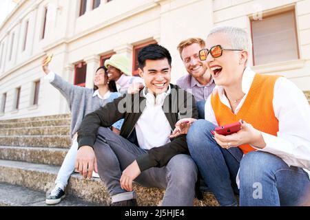 Grande risata di una donna con gli amici che usano cellulare. Gruppo felice di persone che si divertono all'aperto prendendo selfie e sorridendo tenendo smartphone. Foto Stock