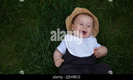 Piccolo bambino neonato felice in estate panama cappello cadere giù posa su erba a piedi nudi in estate Sunny Day. Bambino bambino bambino ragazzo Smacinatura faccia guardare Foto Stock