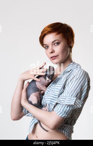 Giovane donna rossa che abbraccia dolcemente il gatto canadese Sphynx, guardando la macchina fotografica. Studio girato su sfondo bianco. Ritratto di hipster con capelli corti Foto Stock