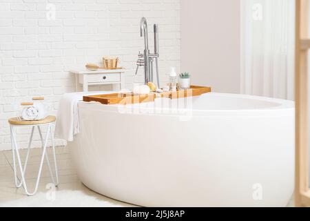 Moderna vasca da bagno con vassoio in legno e diversi accessori nella stanza da bagno interna Foto Stock