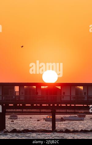 Cielo arancione chiaro con il sole che sorge sopra la silhouette del molo Herne Bar sulla costa del Kent. Dietro e sotto il molo ci sono diverse piccole barche ormeggiate nel porto. Foto Stock