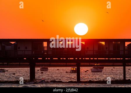 Cielo arancione chiaro con il sole che sorge sopra la silhouette del molo Herne Bar sulla costa del Kent. Dietro e sotto il molo ci sono diverse piccole barche ormeggiate nel porto. Foto Stock